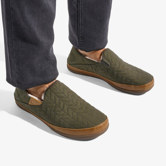 Olive green quilted slip-on slippers with tan rubber sole, worn with dark jeans.