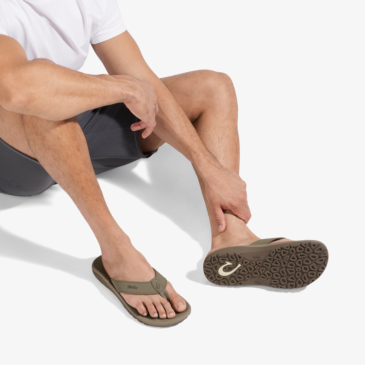 Man wearing white shirt and dark shorts seated, displaying tan thong sandal with textured sole and curved footbed.