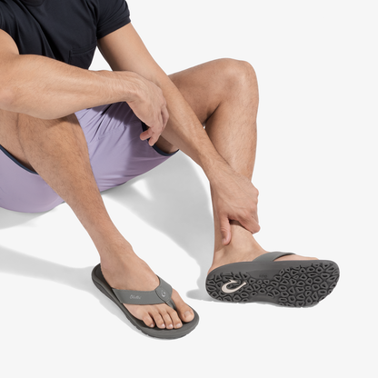 Man wearing dark shirt and lavender shorts seated while wearing gray and black thong sandal with textured sole.