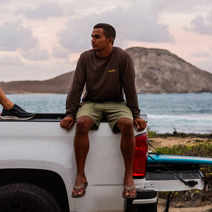 Man wearing dark long-sleeve shirt and olive shorts sitting on white truck bed at beach.