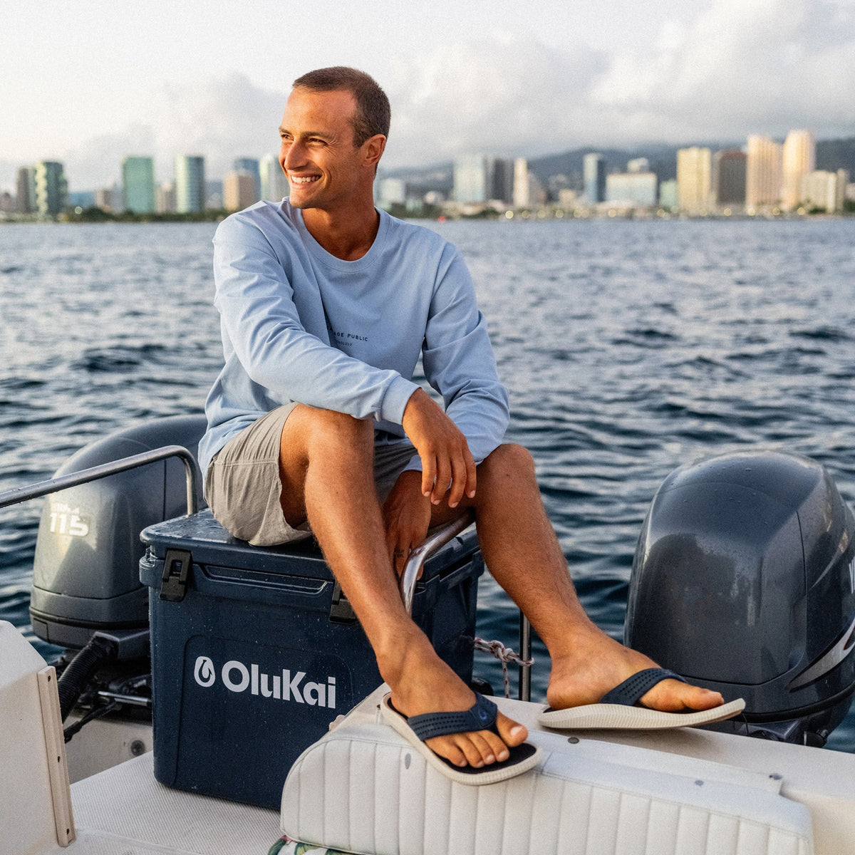 Man wearing light blue long-sleeve shirt and navy slide sandals sitting on boat with city skyline in background.