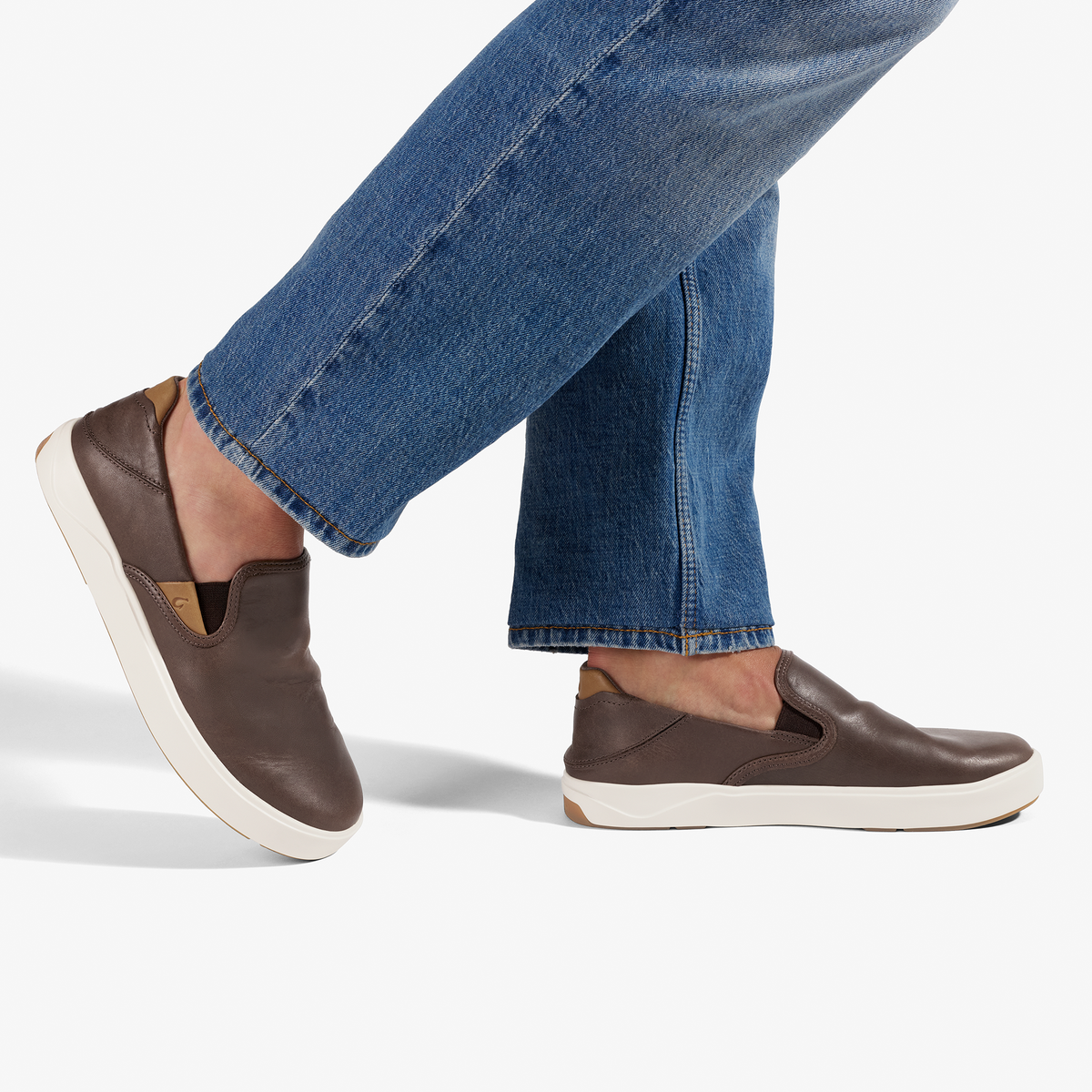 Dark brown leather slip-on sneakers with white sole, worn with blue jeans.