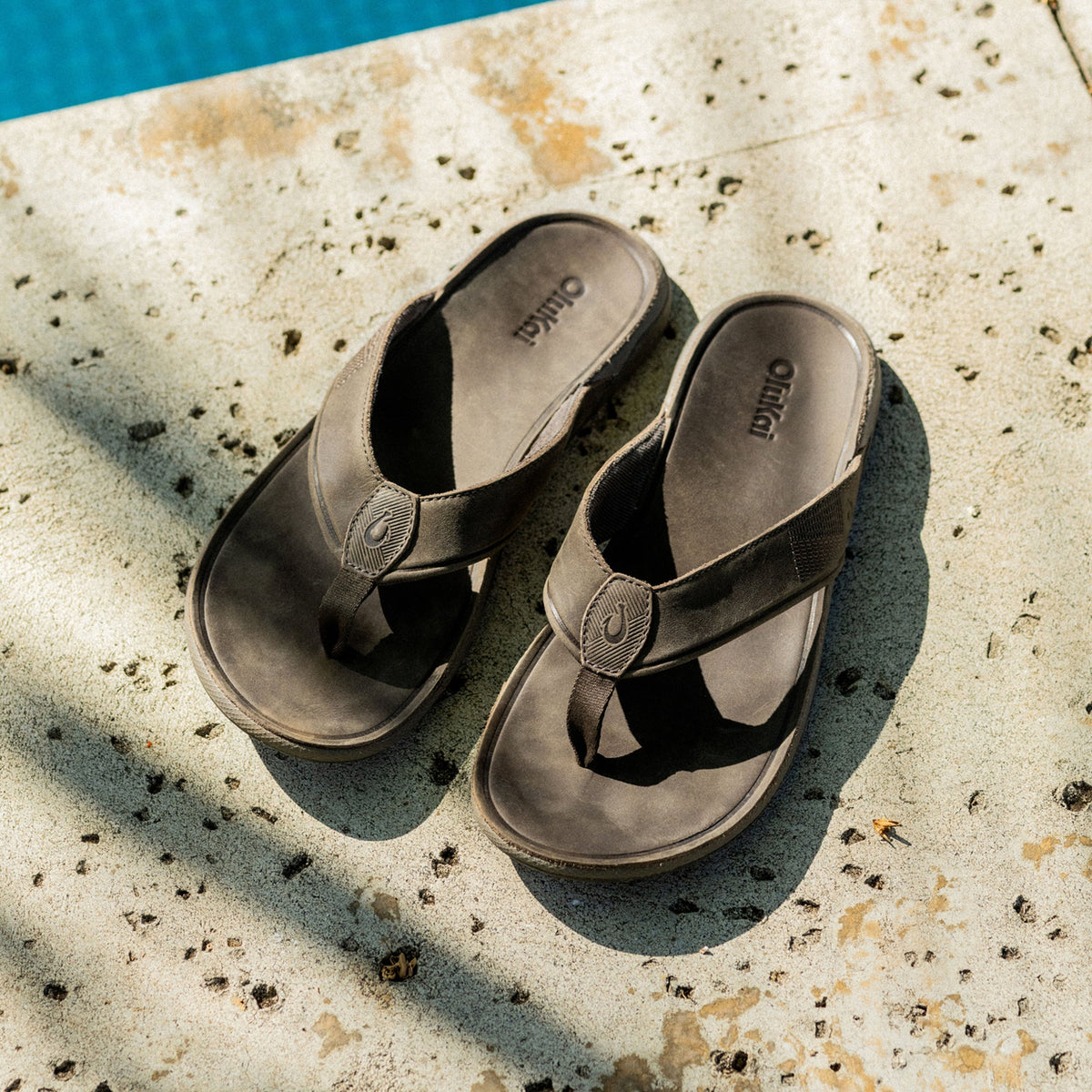 Pair of dark gray thong sandals with embossed circular details on the straps, displayed on a speckled concrete surface near w