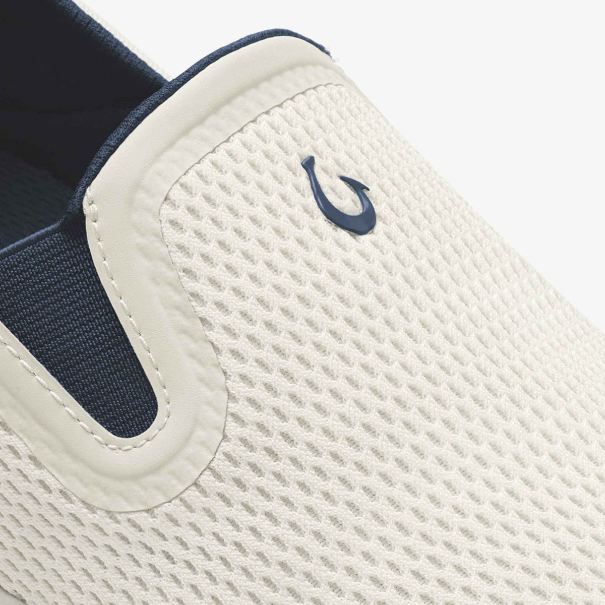 White slip-on shoe with textured mesh upper, navy blue logo on heel, and dark blue elastic side panel.