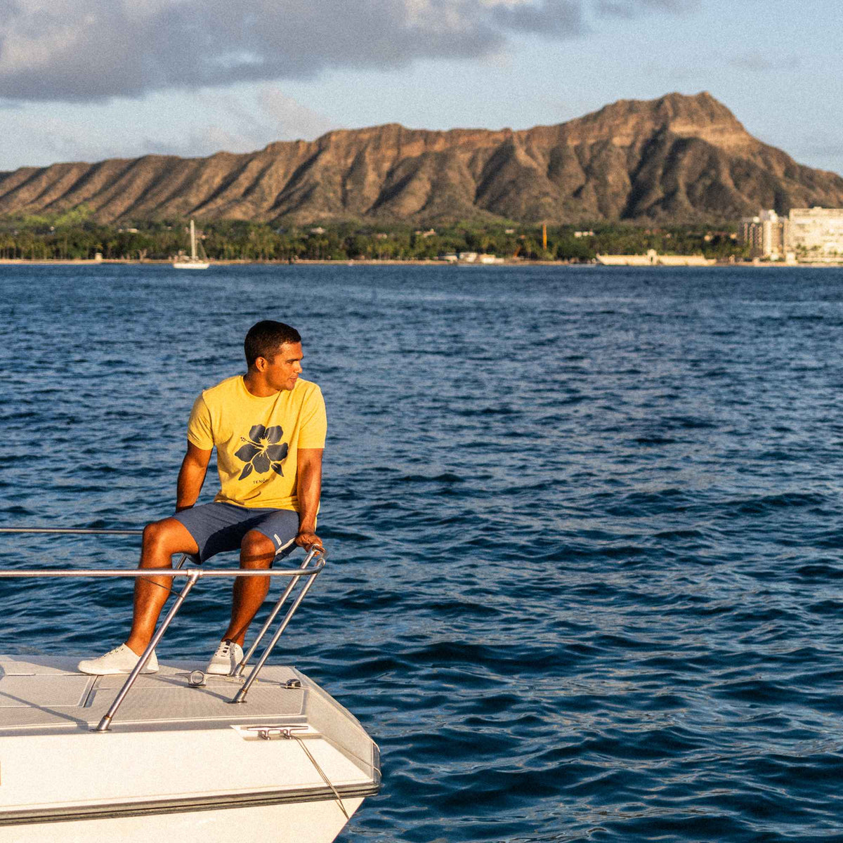 Man wearing yellow graphic tee and navy shorts sits on white boat rail overlooking blue ocean and mountain coastline.
