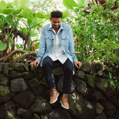 Man wearing light blue shirt, white striped undershirt, dark trousers, and tan canvas shoes sitting on stone wall.