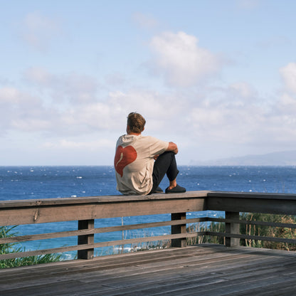 Person wearing tan and red Hāmani Hulu sitting on wooden railing overlooking ocean.