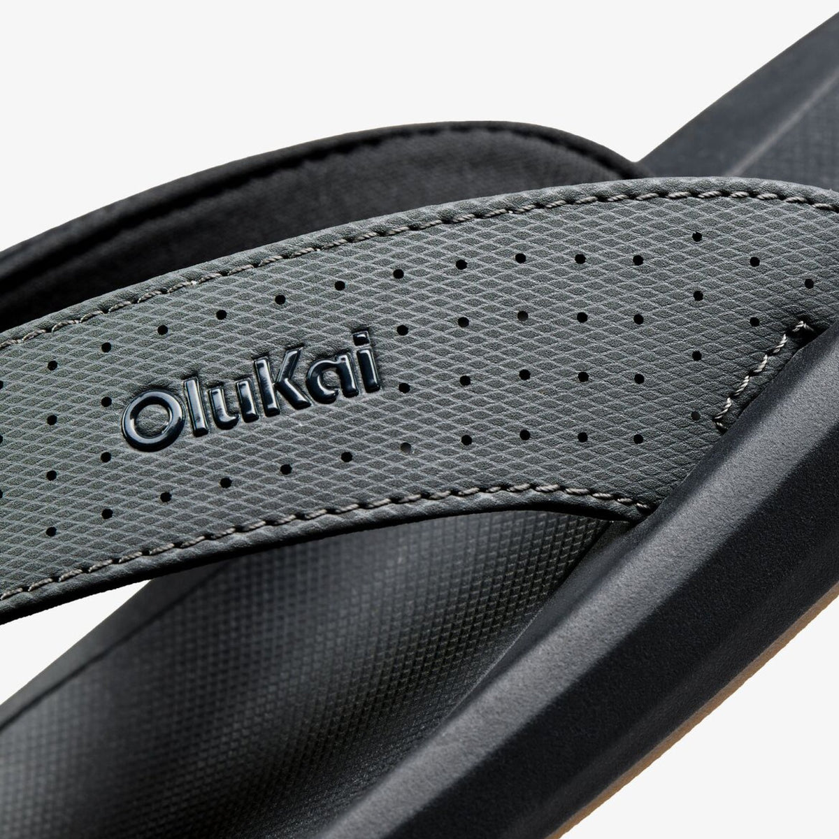 OluKai flip-flop with perforated gray textile strap and black sole, showing brand name and stitching details.