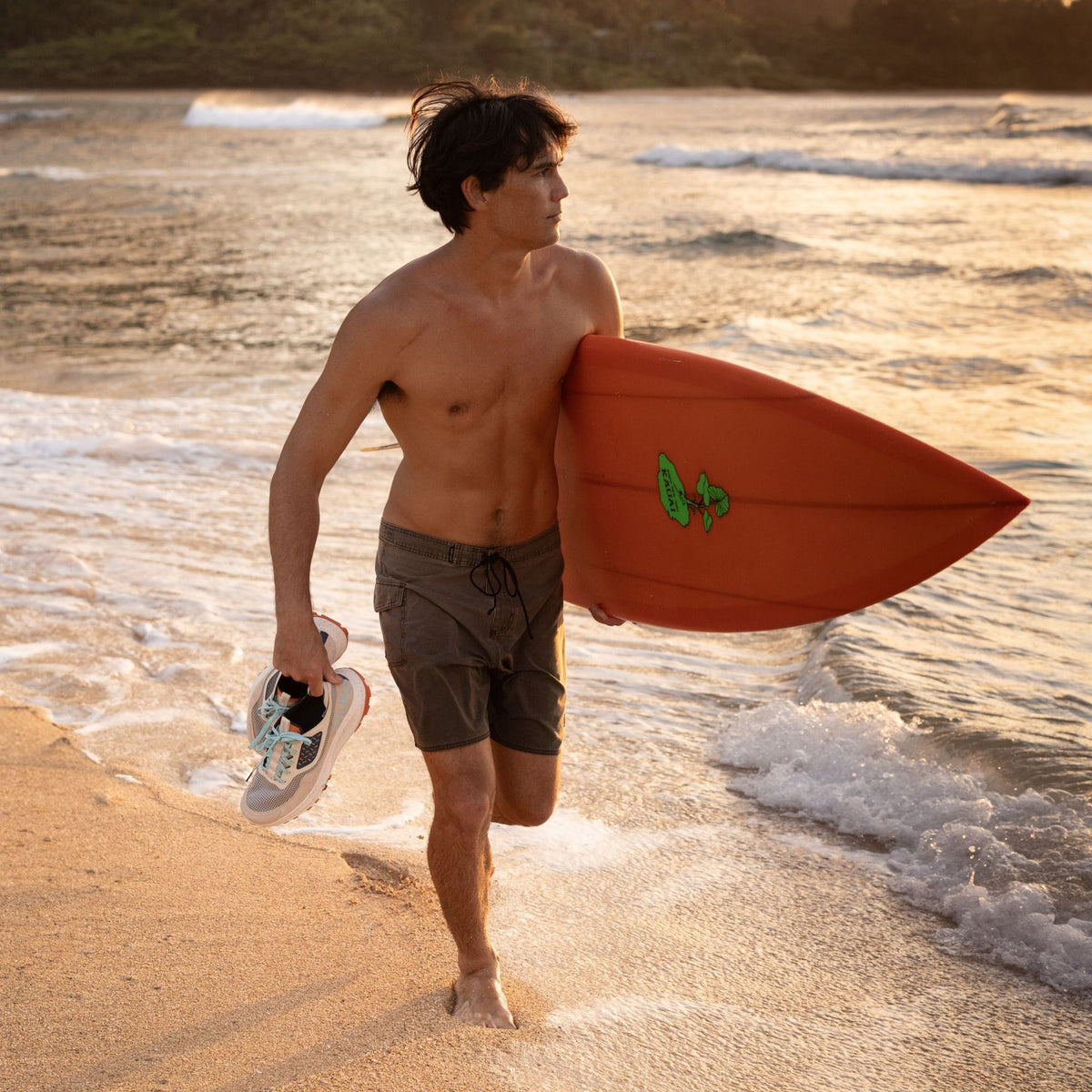 Shirtless man carrying red surfboard with green logo walking on beach toward ocean waves.