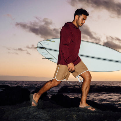 Man in maroon hoodie and tan shorts carrying brown koa surfboard on beach at sunset.