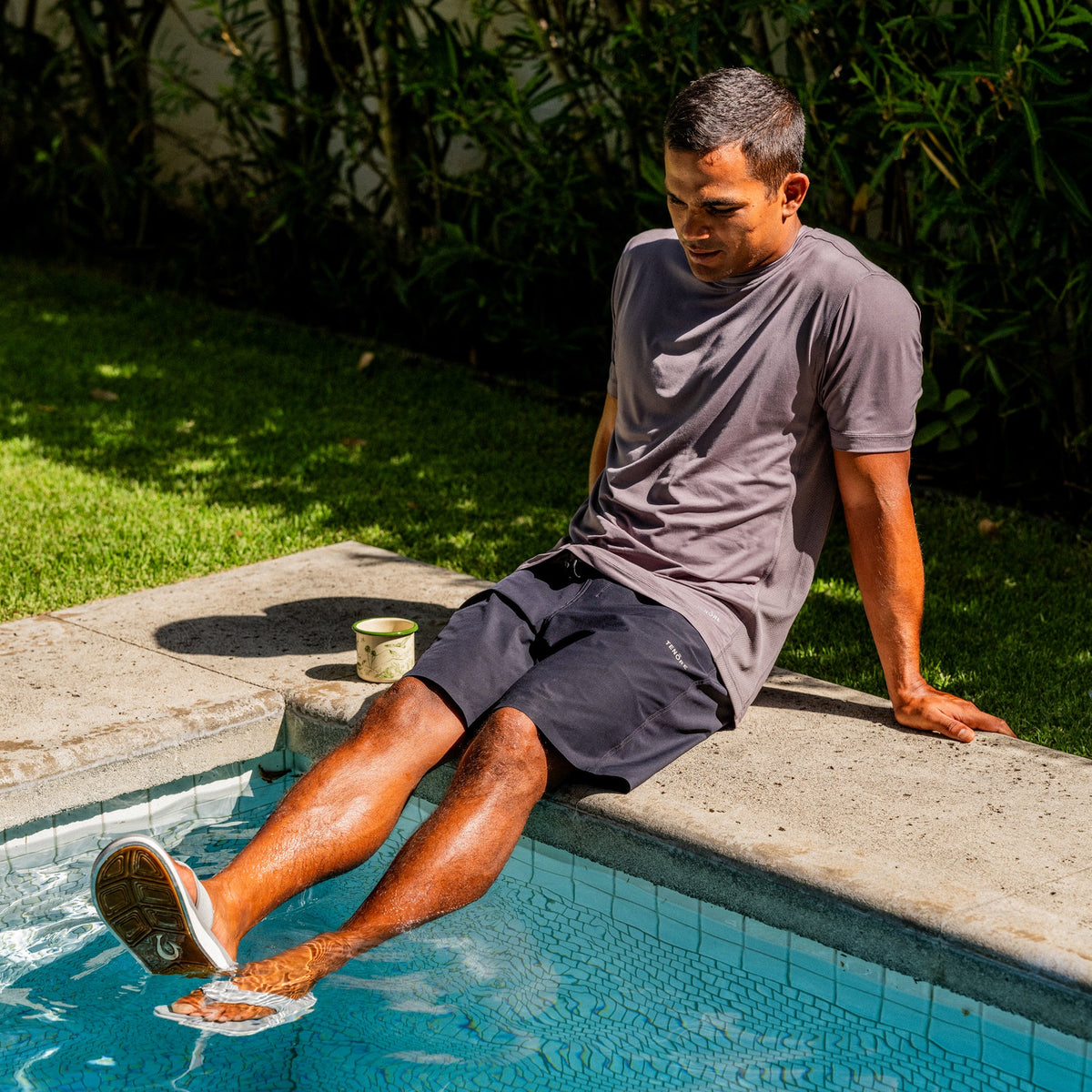 Man in ash gray t-shirt and dark shorts sitting poolside with feet in water.