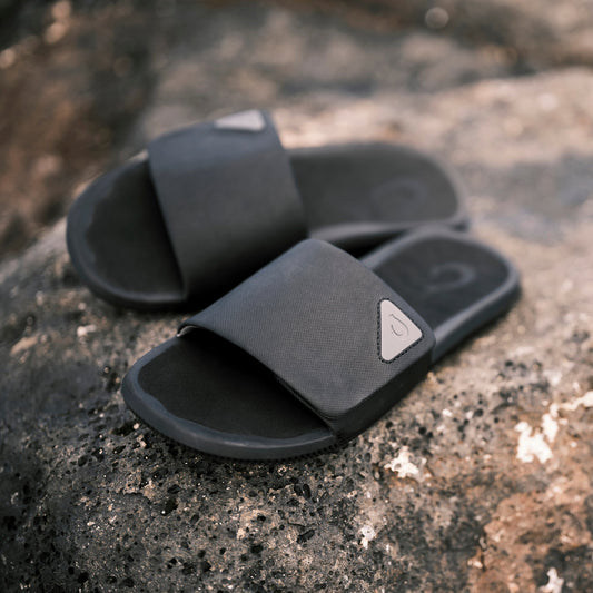 Black slide sandals with textured straps and white logo details on lava rock surface.