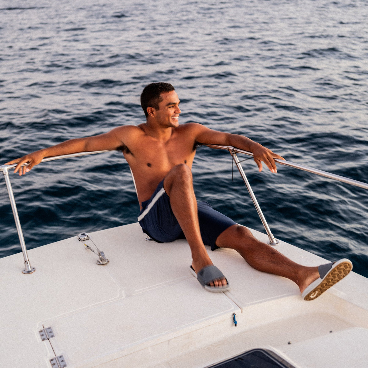 Man wearing navy slide sandals sitting on white boat with ocean in background.