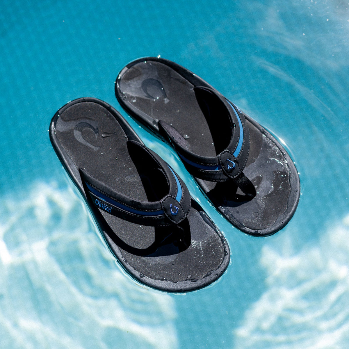 Pair of black and slate blue thong sandals with textured soles, viewed from above on turquoise water.