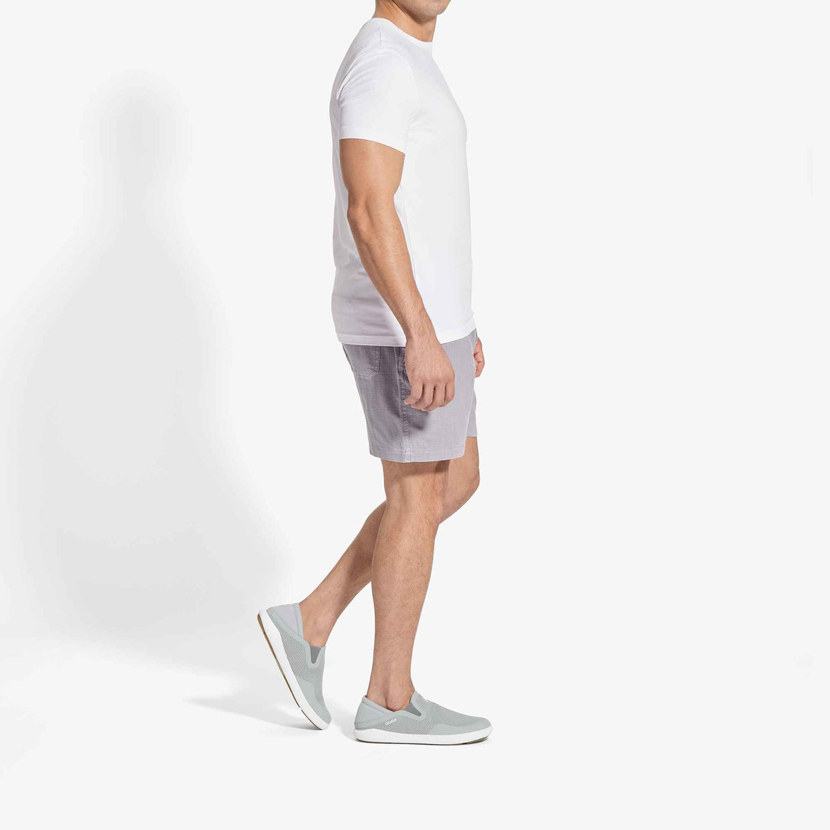 Man wearing white t-shirt, grey shorts, and grey slip-on sneakers shown in side view.