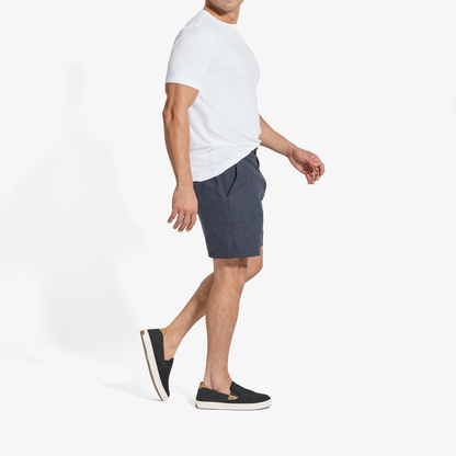 Man wearing white short-sleeve t-shirt, dark blue shorts, and black slip-on sneakers with tan trim.