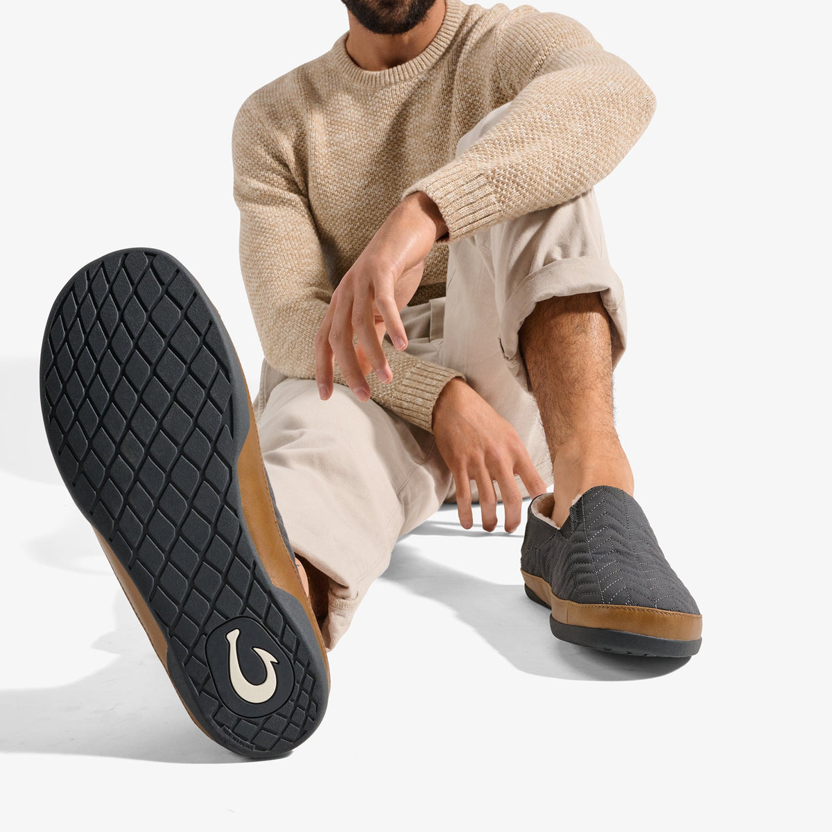 Man wearing beige sweater and cream pants sits displaying dark gray slip-on shoe with quilted sole and tan suede trim.