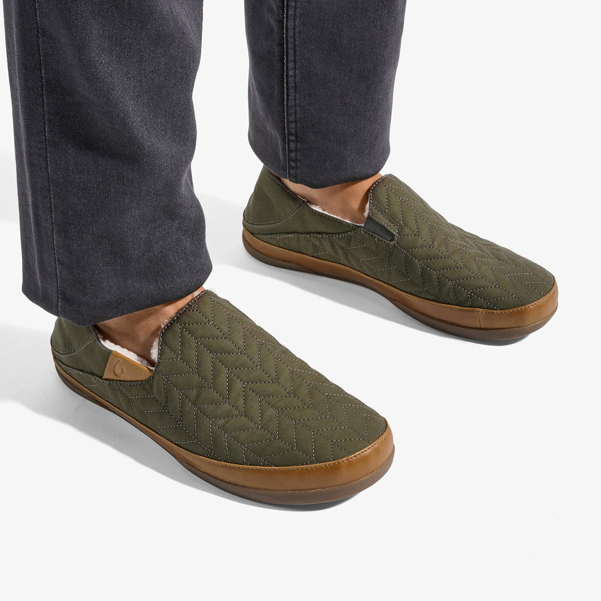 Olive green quilted slip-on slippers with tan rubber sole, worn with dark jeans.
