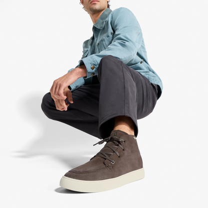 Dark brown suede chukka boot with cream sole, laced closure, worn with black pants and light blue shirt.