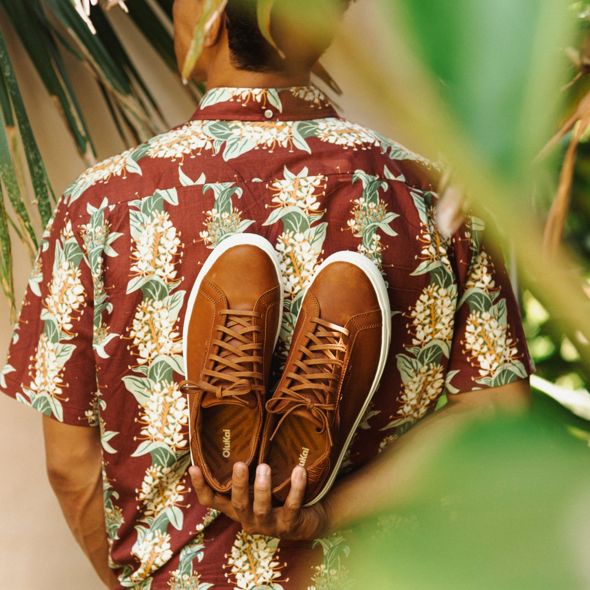 Tan lace-up sneaker with white sole and dark laces, held against a floral burgundy shirt.