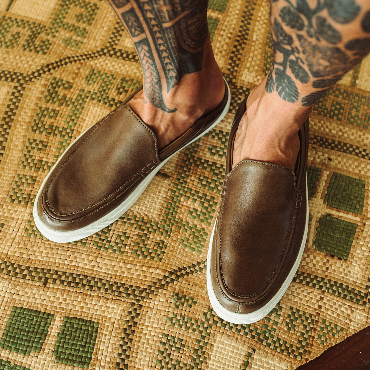 Brown leather slip-on loafers with white rubber soles worn on a patterned yellow and green woven surface.