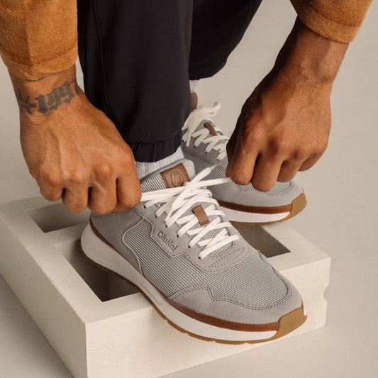 Light grey mesh sneaker with white laces, brown heel tab and sole, worn with black pants.