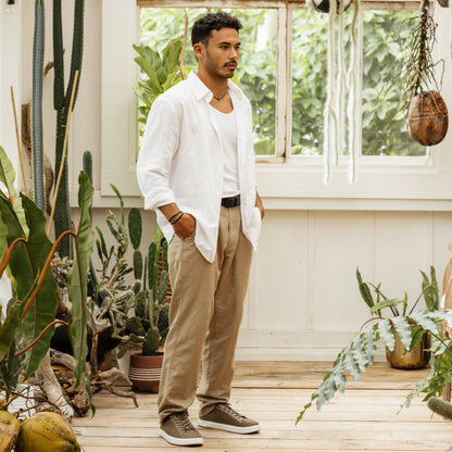 Man wearing white linen shirt and tan pants standing indoors among potted plants and greenery.