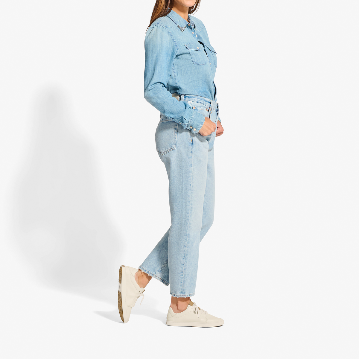 Woman wearing light blue denim shirt and jeans with cream canvas sneakers.