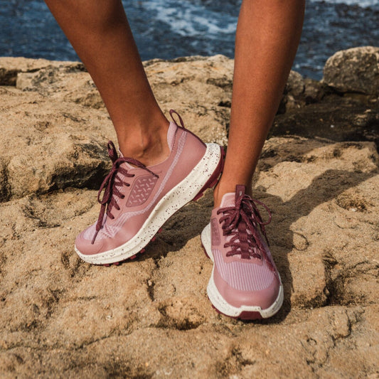 Coral rose lace-up athletic shoes with white soles worn on sandy beach rocks.