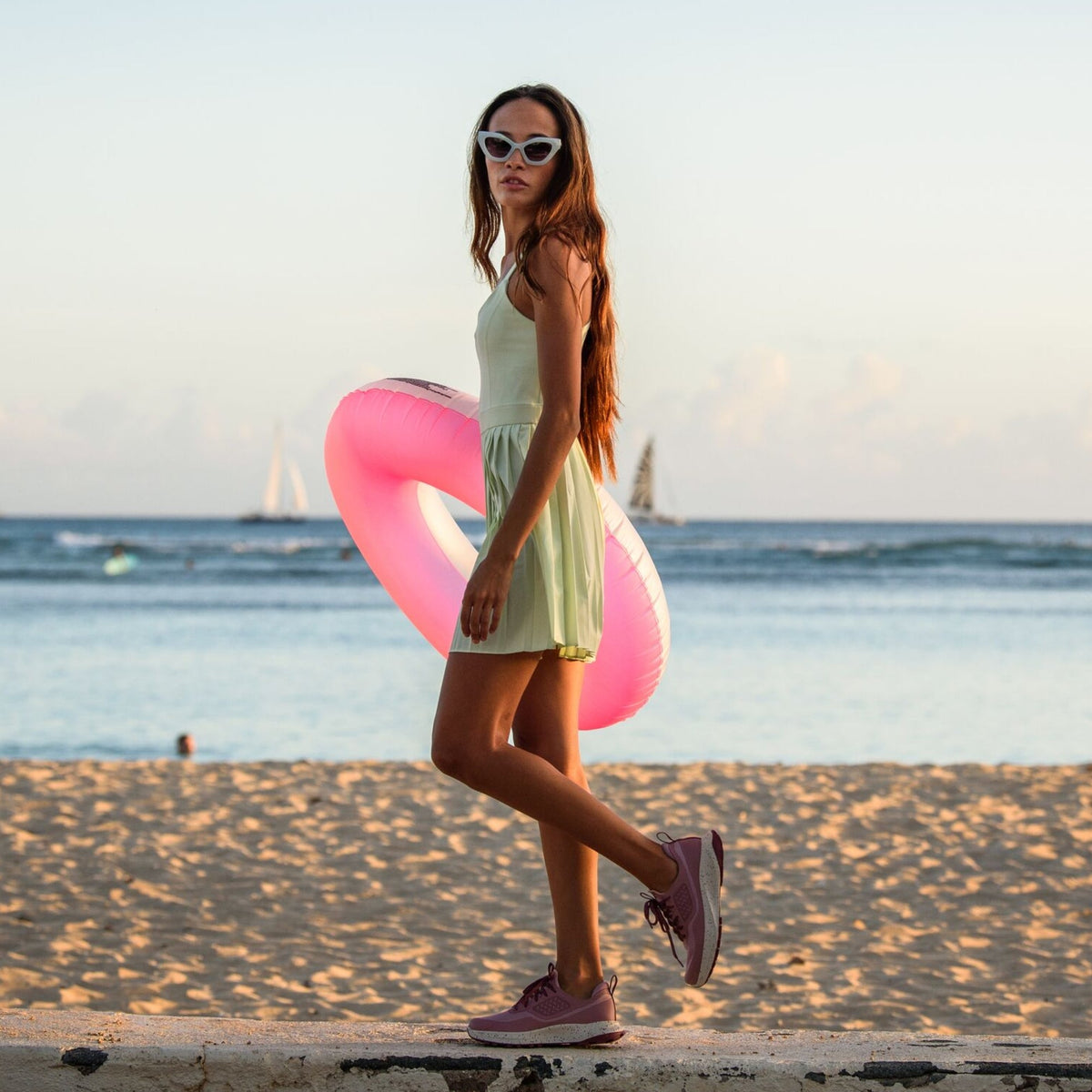 Woman wearing pale yellow sleeveless dress and white sunglasses holding coral pink inflatable ring on beach.