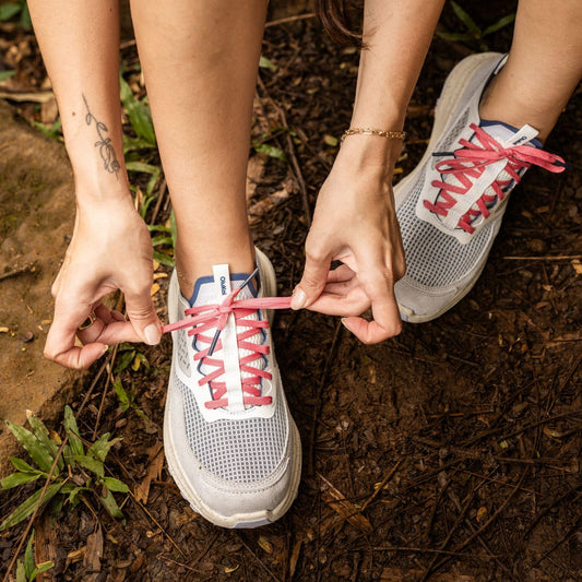 White and blue mesh sneaker with red laces, shown on person's feet outdoors on dirt ground.