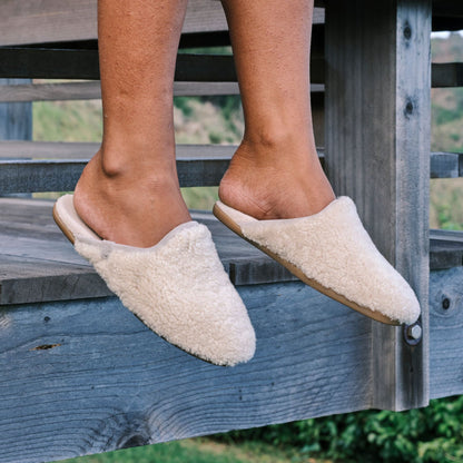 Cream wool mule slippers with tan sole worn by person sitting on wooden bench.
