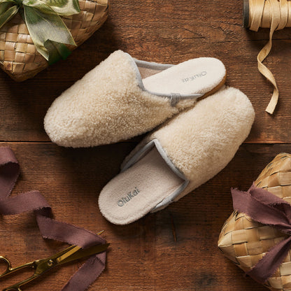 Cream-colored fleece mule slippers with gray trim and text on insoles, displayed on dark wood surface.