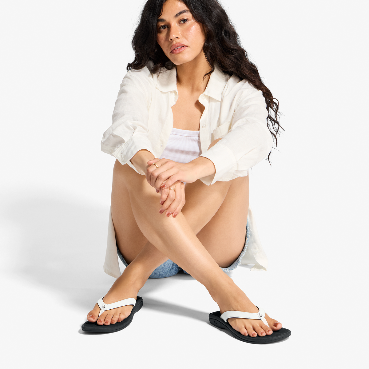 Woman wearing white button-up shirt and white and black flip-flops, sitting with knees bent.