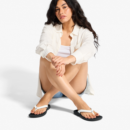 Woman wearing white button-up shirt and white and black flip-flops, sitting with knees bent.
