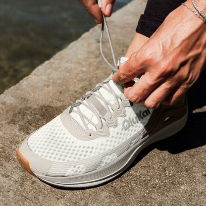 White and gray athletic sneaker with mesh upper and tan sole being held by person's hands near water.
