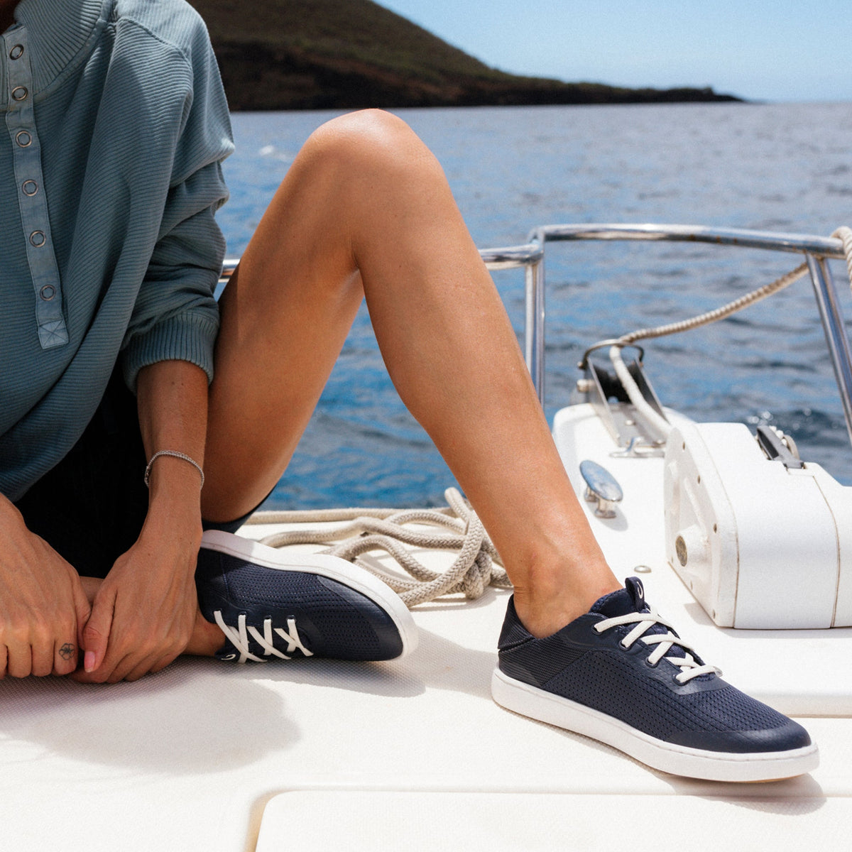 Person wearing trench blue button-up shirt and navy lace-up sneakers with white sole on boat deck.